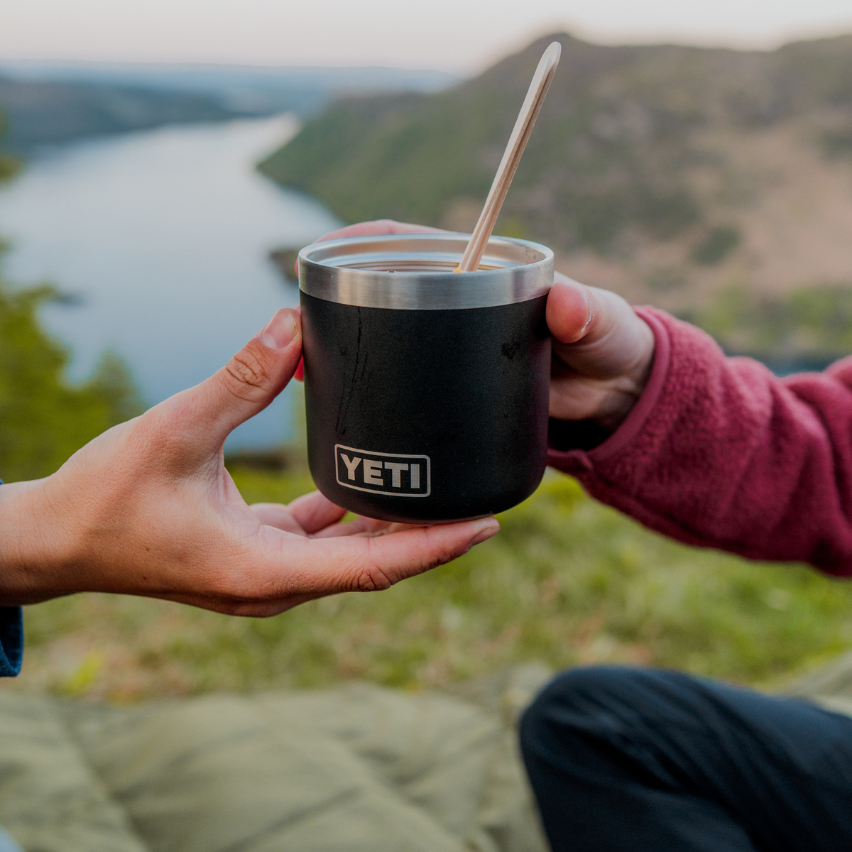 A person outdoors holds a black Yeti Rmabler insulated food jar with a stainless steel rim and a wooden spoon, with a lake and hills blurred in the background.