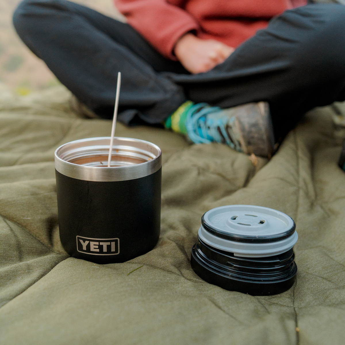 A black Yeti Rambler insulated food jar with a stainless steel rim sits on an olive green blanket outdoors, with a person seated in the background and the food jar lid placed beside it.