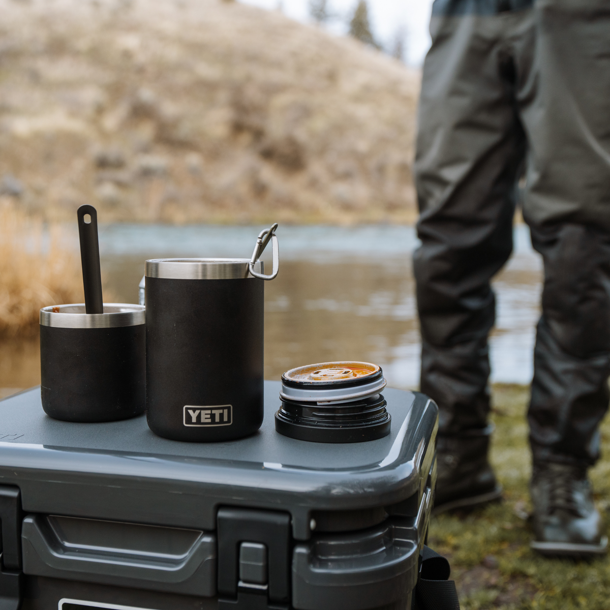 2 black Yeti Rambler Insulated food jars with a stainless steel rims, placed on top of a grey cooler outdoors near a body of water, with a person's legs visible in the background.