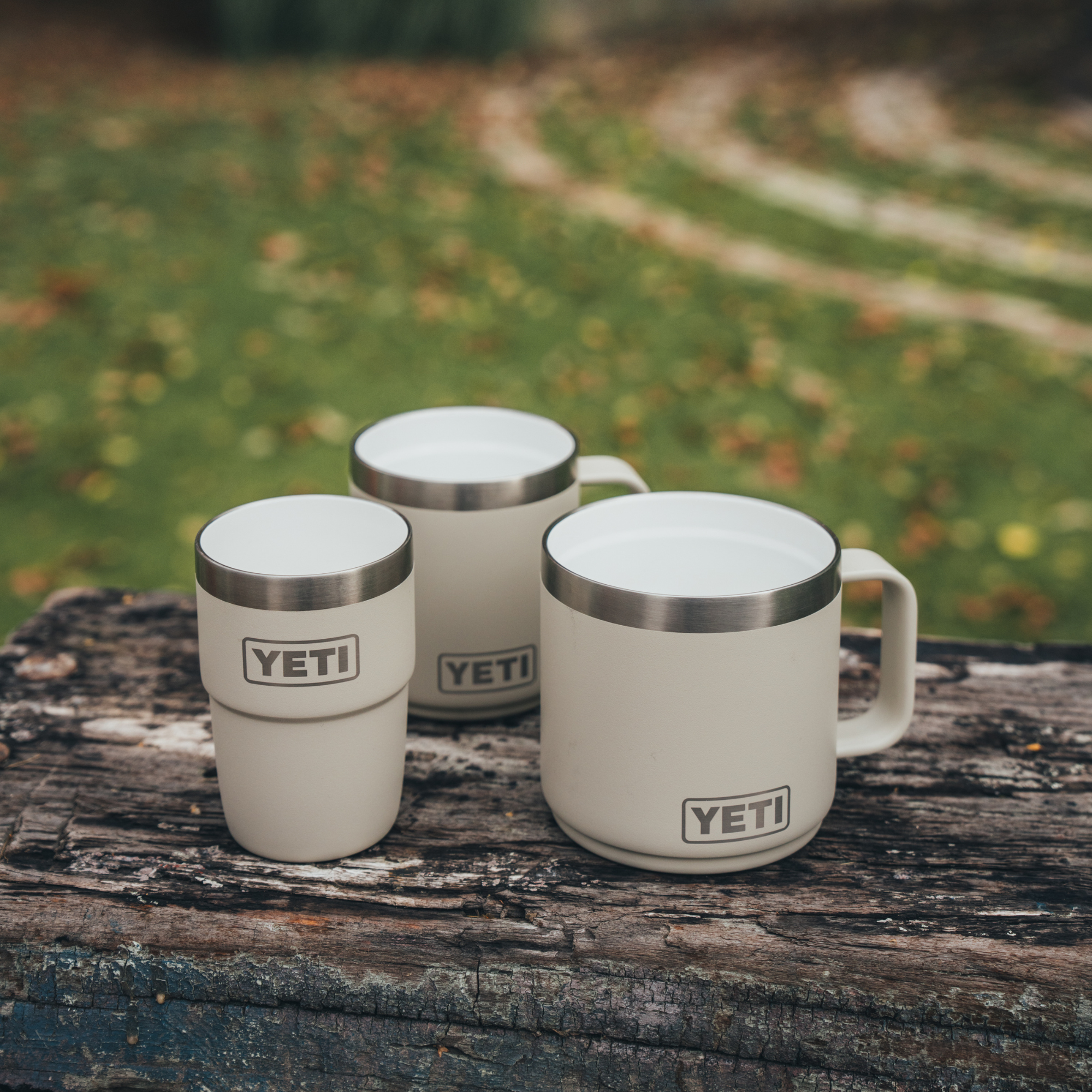 Three cream Cape Taupe Yeti travel mugs, arranged on a weathered wooden log with a blurred outdoor grassy background.