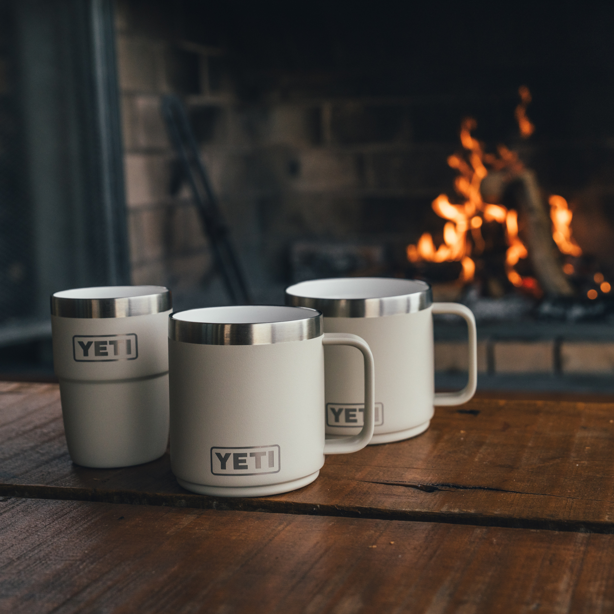Three cream Cape Taupe Yeti travel mugs are arranged on a rustic wooden table in front of a warm, glowing fireplace.