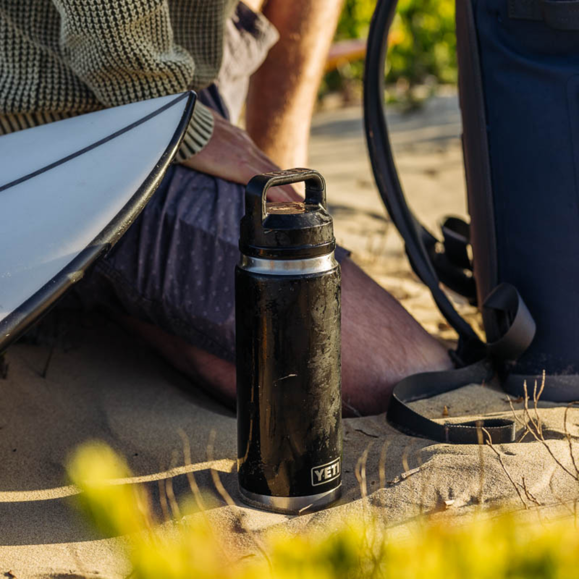 Black Yeti Rambler Insulated Bottle with Chug Cap sitting on the sand beside a man kneeling down beside a surfboard