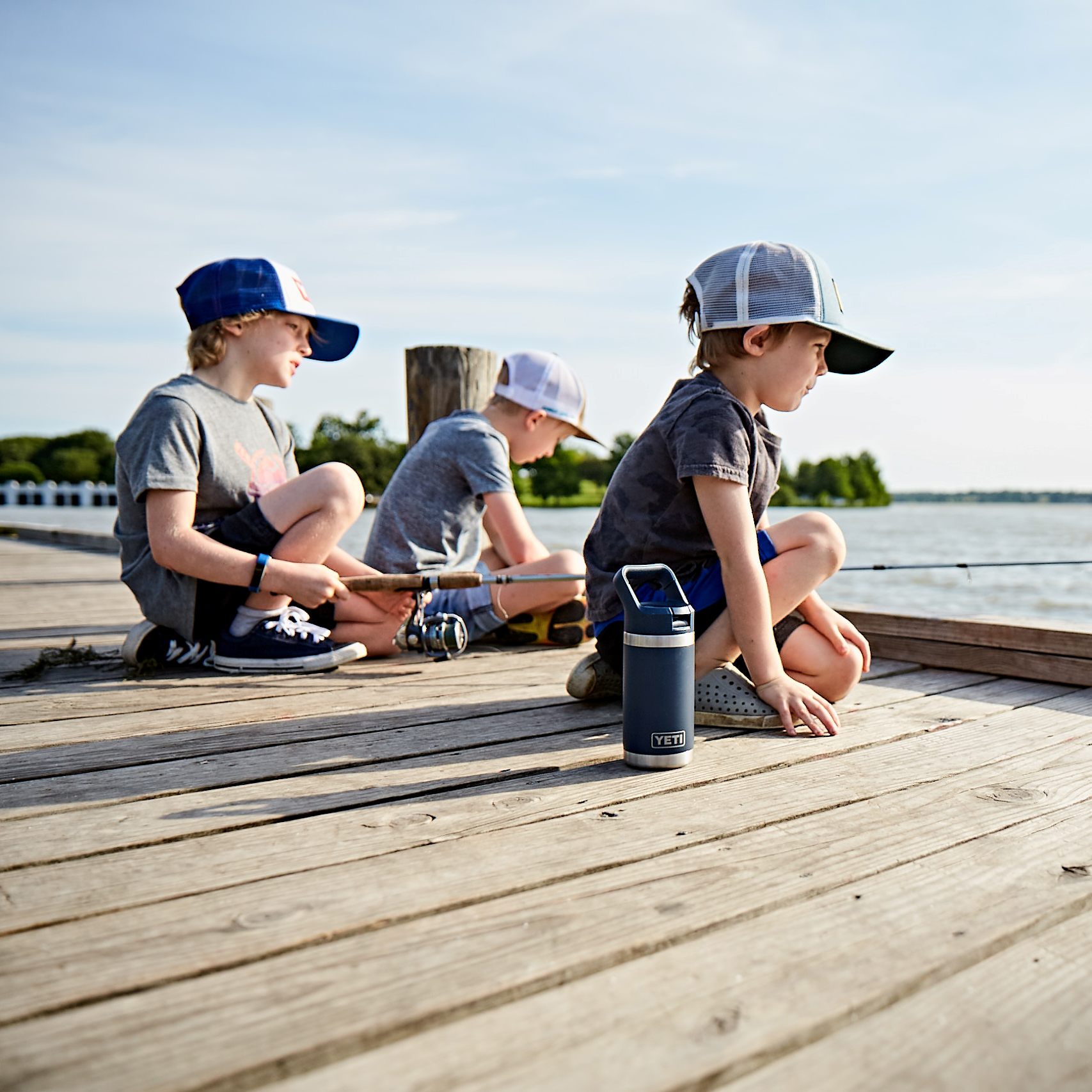 Three children sitting on a wooden dock by a body of water with a Yeti Rambler Junior 12oz 355ml Insulated Bottle with Colour Matched Straw Cap Navy 70000003469 Yeti-RamblerJnr-12oz.