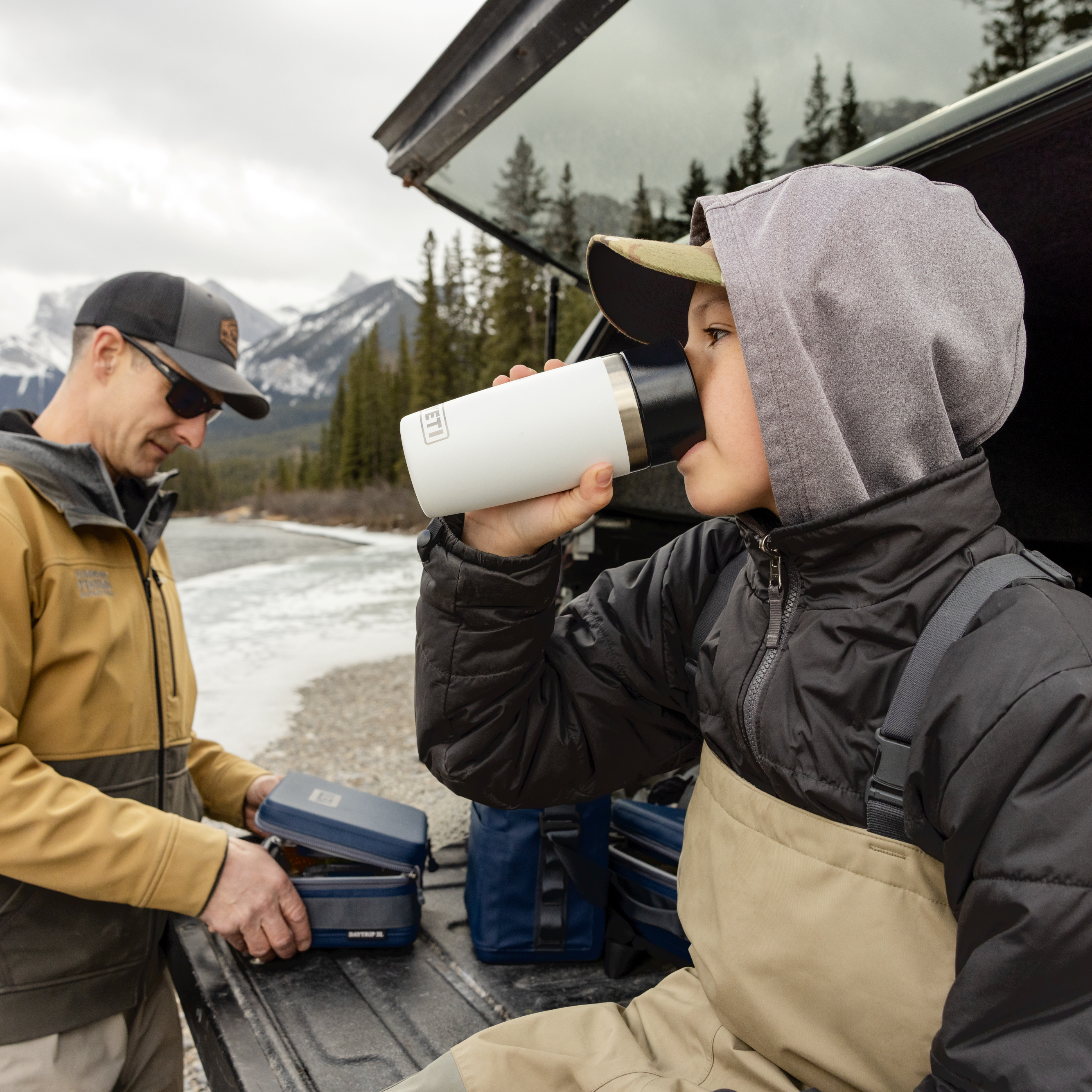 Two people in fishing clothing by a vehicle with mountains in the background drinking out of a Yeti travel mug
