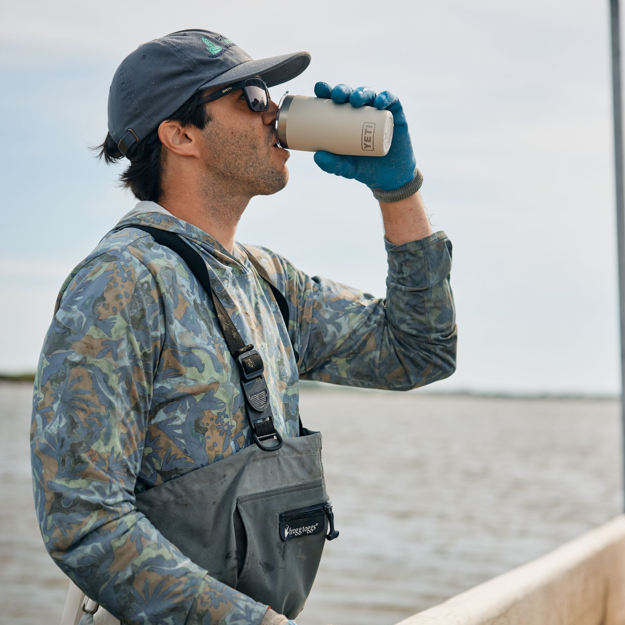 A man wearing a camouflage long-sleeve shirt, grey waders, and a dark cap drinks from a Yeti Rambler Tumbler Insulated Travel Mug Magslider Lid 20oz 591ml Cape Taupe 70000003166 Yeti-RamblerTumbler-20oz on a boat on the water.