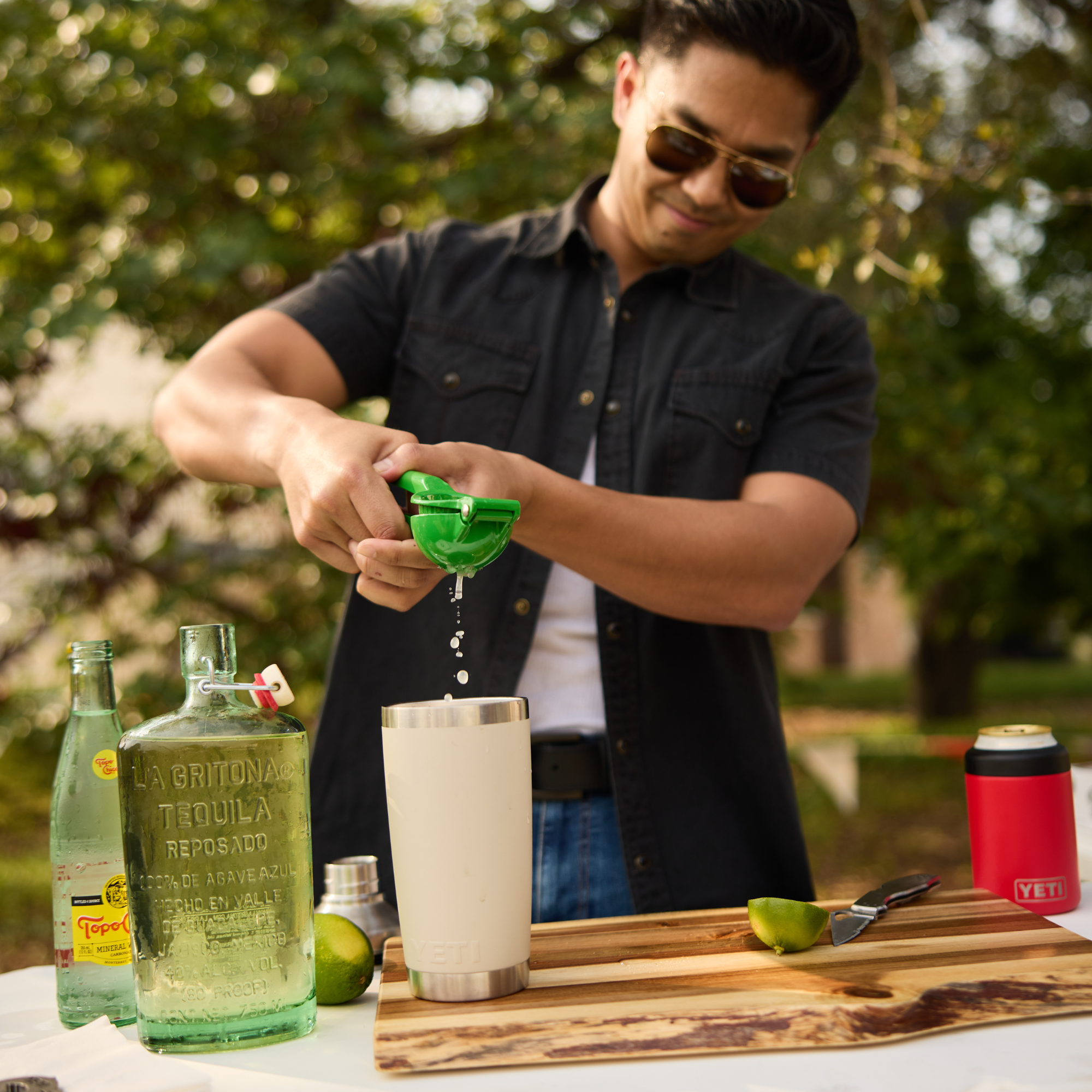 A man wearing sunglasses and a dark button-up shirt squeezes lime juice into a Yeti Rambler Tumbler Insulated Travel Mug Magslider Lid 20oz 591ml Cape Taupe 70000003166 Yeti-RamblerTumbler-20oz over a wooden cutting board, with bottles and limes on a table outdoors.