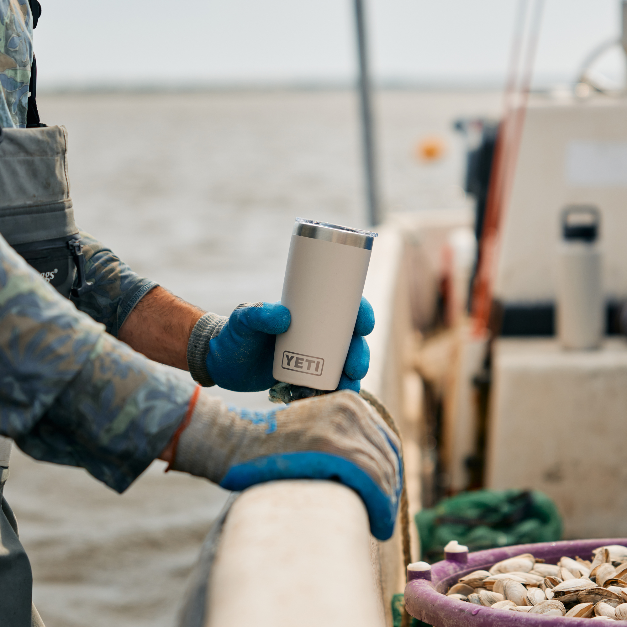 A person on a boat wearing blue gloves holds a Yeti Rambler Tumbler Insulated Travel Mug Magslider Lid 20oz 591ml Cape Taupe 70000003166 Yeti-RamblerTumbler-20oz. The background shows the water and a fishing boat, with a bowl of clams in the foreground.